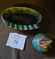 Top-down image showing Green and blue glazed pottery ashtray with raised 'expo67' text and smaller lighter next to it on dark surface.