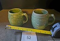 Two green glazed yellowware barrel-shaped beer mugs side by side with measuring tape for scale showing approximately 4 inches in height