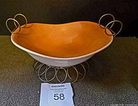Orange pottery bowl displayed in silver-tone metal stand with ring design and loop handles, photographed from the side on dark carpet.