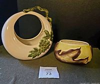 Two vintage ceramic vases side by side on a dark background, showing Hull Tokay circular vase with grape embossing and Royal Copley planter with green and brown glaze.