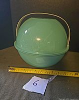 Closed aqua plastic ball container with handle snapped in place, shows size relative to tape measure, approx 10.5 inch diameter