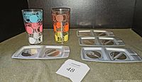 Two 1960s highball glasses with colorful circle patterns and four aluminum divided trays for snacks and cigarettes on display.