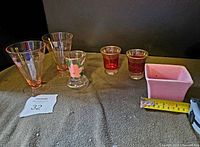 Lot overview showing all glassware and ceramic planter including two pink etched parfait glasses, footed painted glass, two small etched shot glasses, two small pink flower vases, and pink ceramic planter with measuring tape.