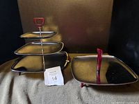 3-tier chrome serving stand with cherry red Bakelite handles next to a flat serving tray with matching handle and feet, photographed on a dark surface.
