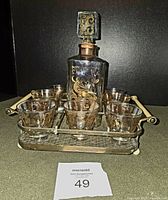 Vintage midcentury decanter with gold floral motifs alongside 8 matching shot glasses on a metal tray with white handles