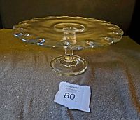 Side angle of clear glass pedestal cake stand with scalloped edge and teardrop indentations on plate.