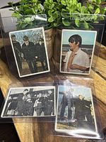 Four Beatles trading cards from 1964 O-Pee-Chee, featuring color and black-and-white images of the band, placed on a wooden surface with green plant background.