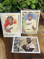 Three Jackie Robinson Dodgers baseball cards from Topps 1997, Topps Allen & Ginter 2020, and Topps Ginter 2023 displayed upright against a green plant background on wooden surface.