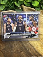 Photo of limited print 2024 Topps Now USA Basketball trading card featuring Curry (#4), LeBron James (#6), and Durant (#7). Card commemorates gold medal win at Olympics.