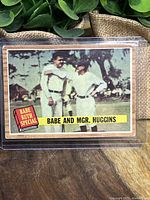 Front view of the vintage 1962 Topps Babe Ruth Special card #137 featuring Babe Ruth and Manager Huggins, set against a baseball field background, protected in a clear plastic sleeve.