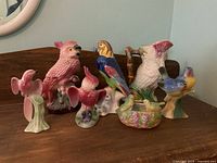 Wide shot showing multiple porcelain bird figurines including pink cockatoos, a parrot, a bird-shaped pitcher, and small birds on bases.