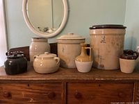 Seven ceramic pots and crocks arranged on a wooden surface with a mirror on the wall behind them, showing different sizes and glaze colors