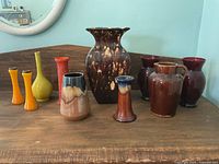 View of 10 assorted ceramic vases of various colors and shapes arranged on a wooden surface against a light blue wall, showing size and finish variety