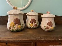Set of three ceramic mushroom-themed cookie jars with lids on wooden surface against teal wall.