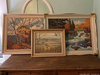 Image showing all three framed paintings arranged side by side on a wooden surface against a wall.