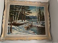 Framed landscape painting of a winter scene with trees and a bridge over a river, with mountains in the distance. Frame has ornate cream-colored detailing with visible scratches and some dust.