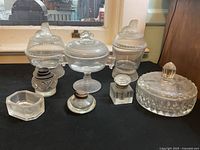 Front view of the seven glassware items including covered dishes, ink bottles, and a rectangular dish on a black surface with a window reflection in the background.