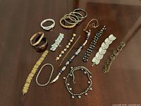 Overview of 14 assorted bracelets and one necklace laid on wood surface. Variety of metal, beaded, and textured styles.