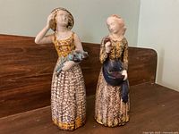 Two ceramic figurines of standing women displayed on a wooden surface against a wall corner, showing the overall height and detail of each piece.