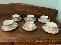 Five Mintons porcelain tea cups with matching saucers arranged on a wooden surface, decorated with pink rose and blue ribbon patterns with gold trim.