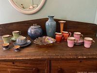 Wide shot of assorted ceramic and porcelain china items on wooden surface including vases, teacups, sugar bowl, and decorative dishes showing colors and condition.