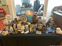 Wide view of owl collection on black tablecloth showing multiple owl figurines in various materials including ceramic, wood, metal, and fabric pillow.