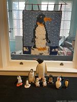 Photo showing various penguin figurines and the latch hook wall hanging hung on a window; sunlight and background visible through window.