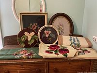 Photo showing multiple framed needlepoint floral art pieces, embroidered cloths, and a hooked rug arranged on a wooden surface.