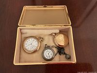 Three pocket watches in a box: two gold-tone watches and one silver-tone watch on chain with fobs