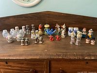 Front view showing entire assortment of shakers arranged on wooden surface including glass, ceramic, and metal types in different designs and colors.