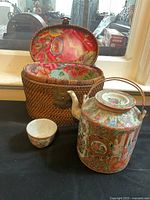 Porcelain tea pot, tea cup, and woven basket carrier displayed together on a table.