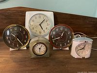 Five assorted clocks displayed on a wooden surface: two black face electric clocks with gold trim, one white square clock, one small metal desk clock, and one white quartz travel alarm clock.