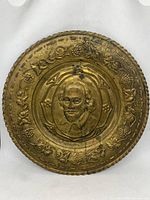 Front view of circular gold coloured wall plate with embossed portrait of William Shakespeare and decorative grapevine border.