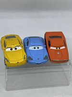 Front view of three colorful Disney Pixar Cars diecast toy cars on a transparent stand