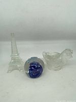 Three glassware items including clear glass small Eiffel Tower figurine, blue glass paperweight with bubbles, and clear glass bird figurine.