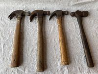 Four hammers including three claw hammers and one ball peen hammer laid on white cloth, showing wooden handles with signs of wear and metal heads with moderate rust.