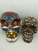 Two skull sculptures side by side on a white background; one is colorful with floral patterns, the other is bronze colored made of multiple smaller skulls.
