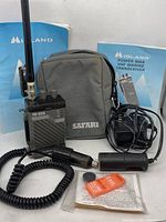 Black Midland VHF Marine handheld CB radio model 78-205 with antenna standing upright beside gray Safari brand soft carrying pouch, two Midland manuals, coiled connecting cord, car lighter outlet power adapter, and orange emergency whistle in packaging.
