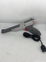Photo showing the Nintendo NES gray Zapper with visible red trigger, attached cable and connector, placed on a white background horizontally.
