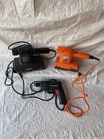 Three tools arranged side by side: two palm sanders (one black, one orange) and one black corded drill with orange cord, on a white fabric background.