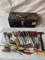 Photo showing a black metal toolbox and an assortment of various hand tools including screwdrivers, pliers, scissors, hammer, and box cutter laid out on a fabric surface.