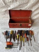 Red metal toolbox open with assorted hand tools laid out in front, including screwdrivers, pliers, wire cutters, various handles in red, yellow, orange, blue, and black.