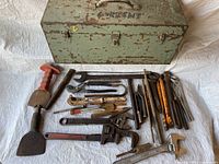 Photo showing vintage metal toolbox and assortment of vintage hand tools including wrenches, pliers, chisels, and files on a light surface.