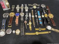 Photo of assortment of 31 various used wristwatches and pocket watches laid out on black background. Shows different styles and bands including metal, leather, fabric, and chain-link. Mixed men's and women's designs.