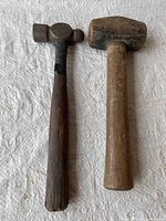 Two vintage hammers displayed side by side on a white textured surface: a small sledge hammer with a light wooden handle and a metal head, and a ball peen hammer with a dark wood handle and metal head partially wrapped with black tape.