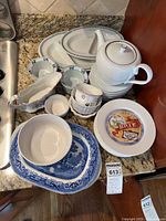 Photo showing white Tiffany palladium band teapot, multiple blue willow pattern platters, various small ramekins, white divided dishes, and appetizer plates with cheese-themed designs stacked on granite countertop.