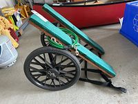 Rolling plank dolly folded showing green padded foam supports and one visible large black plastic wheel with a spoked design.