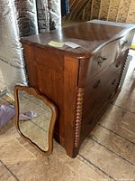 Antique wooden dresser with four drawers, decorative side detail, and detached mirror leaning next to it on the floor.