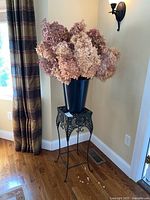 Full view of black metal plant stand holding black plastic pail with large dried hydrangea bouquet inside near window and curtain