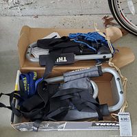 Folded kayak roof rack components with straps and ropes in an open cardboard box on concrete ground.